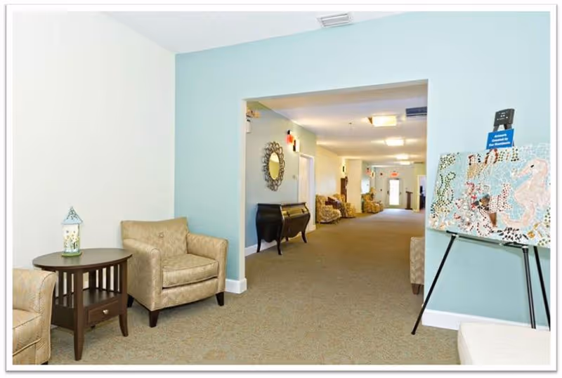 Bright senior living common hallway with upholstered chairs, a side table, decorative mirror and artwork on an easel.