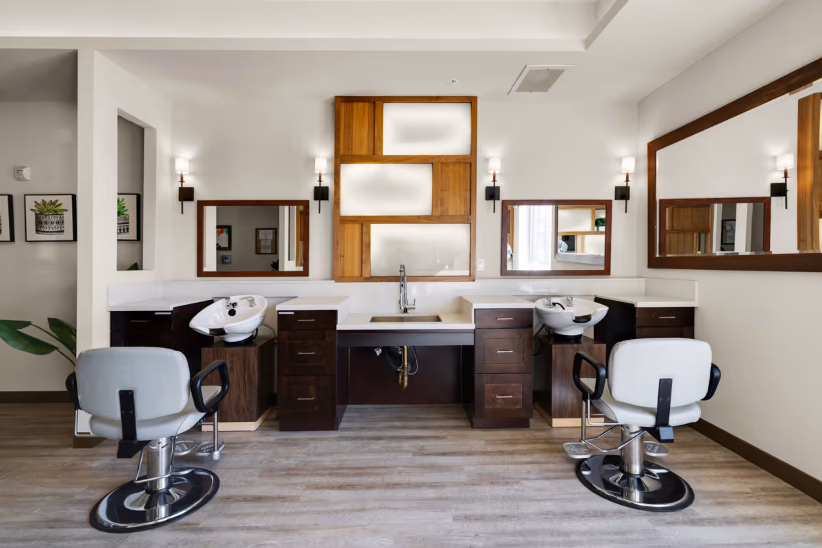 Interior view of a senior living facility's hair salon area featuring two salon chairs in front of wash basins with mirrors on the wall and wooden cabinetry under the counter.