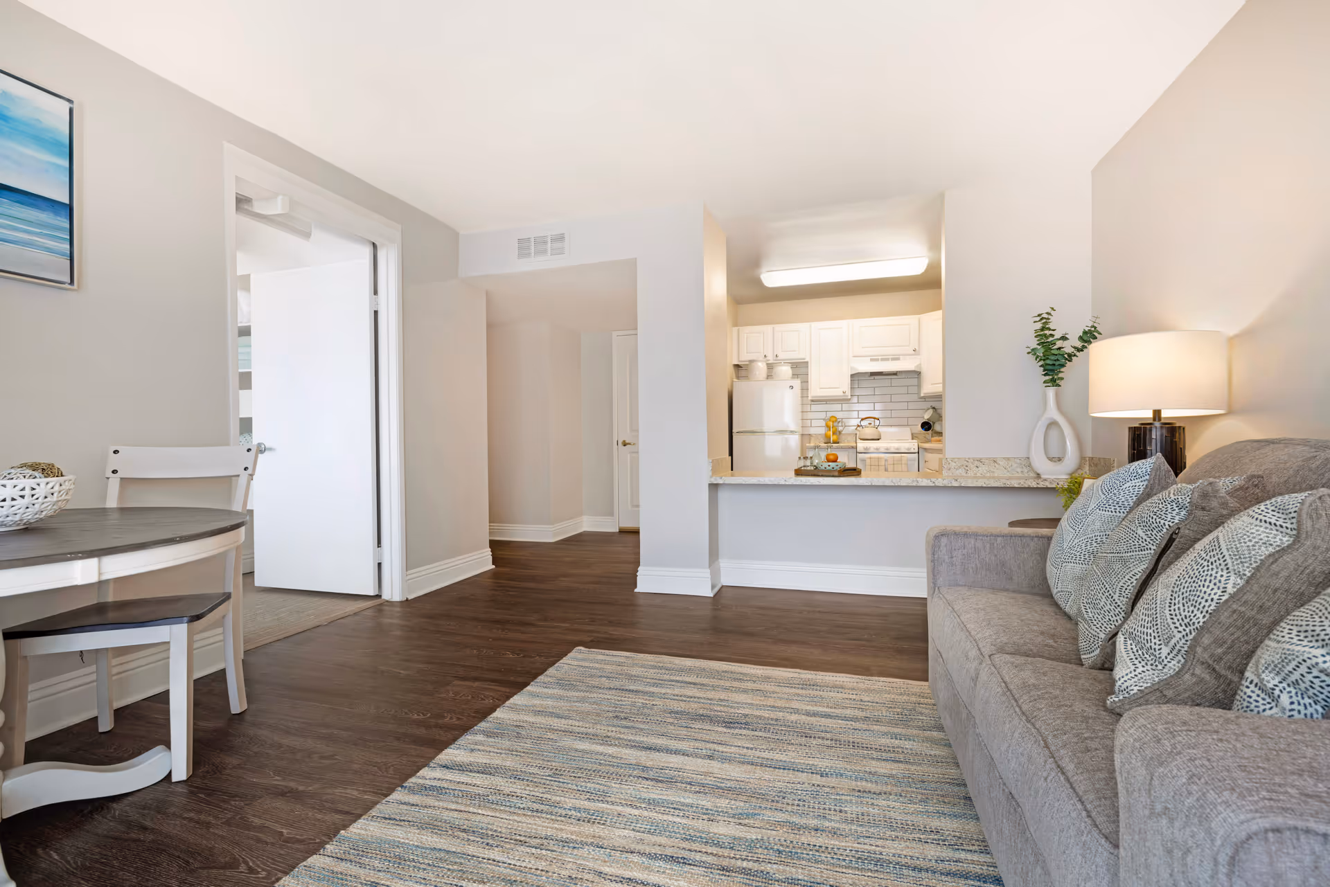 A cozy living room area with a gray sofa adorned with patterned pillows, a table lamp on a side table, a striped area rug on dark wood flooring, and a small round dining table with two chairs. The room opens into a kitchen with white cabinets, a refrigerator, and a countertop with decorative items. A hallway and an open door leading to another room are visible in the background.