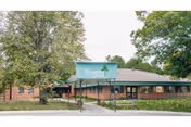 Exterior view of a single-story brick building surrounded by trees and greenery. A sign in front of the building reads 'Goshen Healthcare Community'. The sky is overcast.