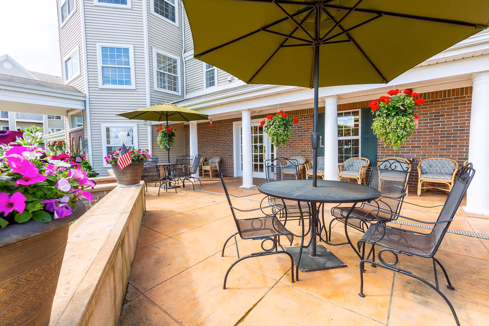 Outdoor patio area at Independence Village Of Petoskey featuring metal tables with green umbrellas, metal chairs, hanging flower baskets with red flowers, potted flowers with American flags, and wicker seating against a brick wall with white columns.