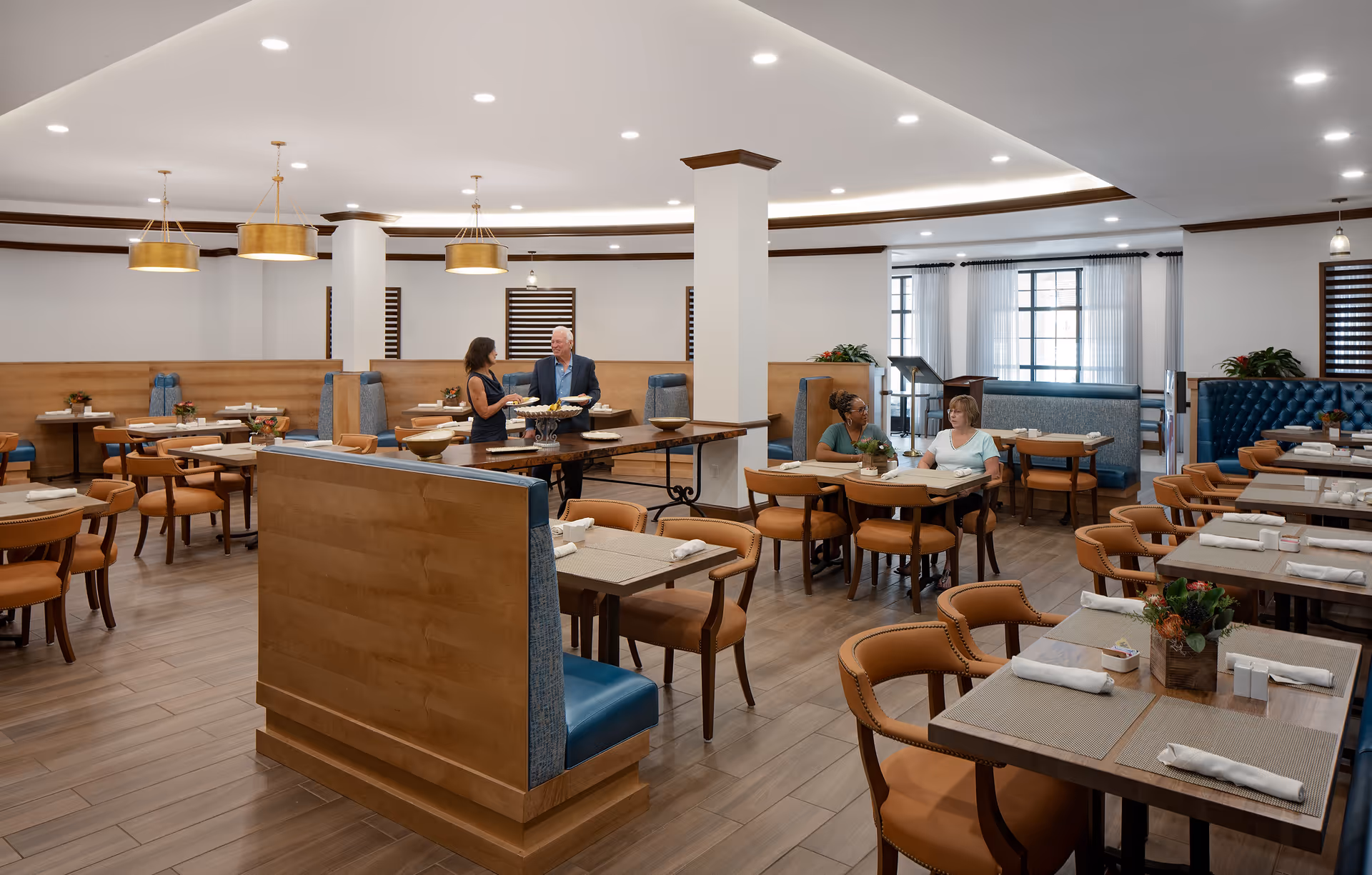 Spacious dining room with tables, booths, pendant lights, and several residents seated and talking.