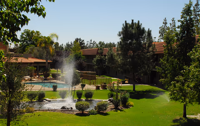 Outdoor view of a senior living facility with a well-maintained green lawn, trees, a small pond with a water fountain, and a fenced swimming pool. The building with a red-tiled roof surrounds the garden area under a clear blue sky.
