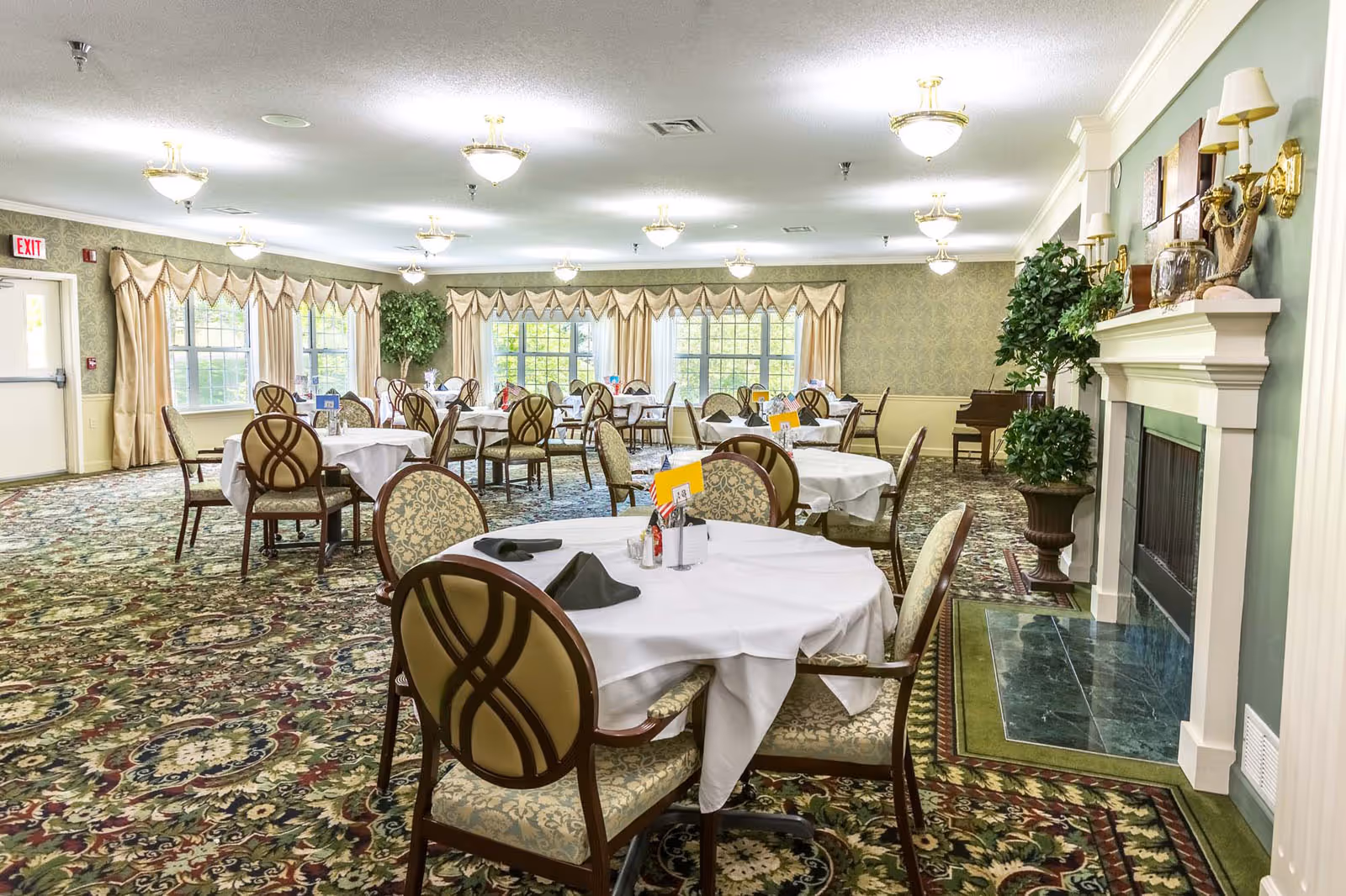 A spacious dining room with multiple round tables covered with white tablecloths and set with black napkins. The room features patterned carpet, large windows with beige curtains, a fireplace with decorative items on the mantel, and a piano in the corner. The lighting consists of multiple ceiling-mounted chandeliers.