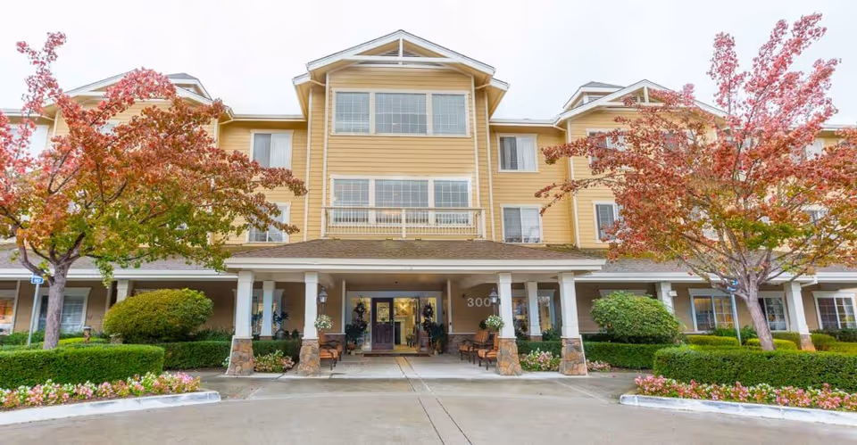 Front exterior view of a three-story senior living facility building with a covered entrance, surrounded by landscaped bushes, flowers, and trees with autumn-colored leaves.