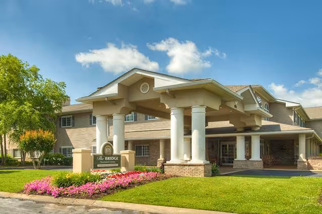 Front entrance of The Bridge at Columbia assisted living building with a covered portico, white columns, manicured lawn and flowerbeds under a blue sky.