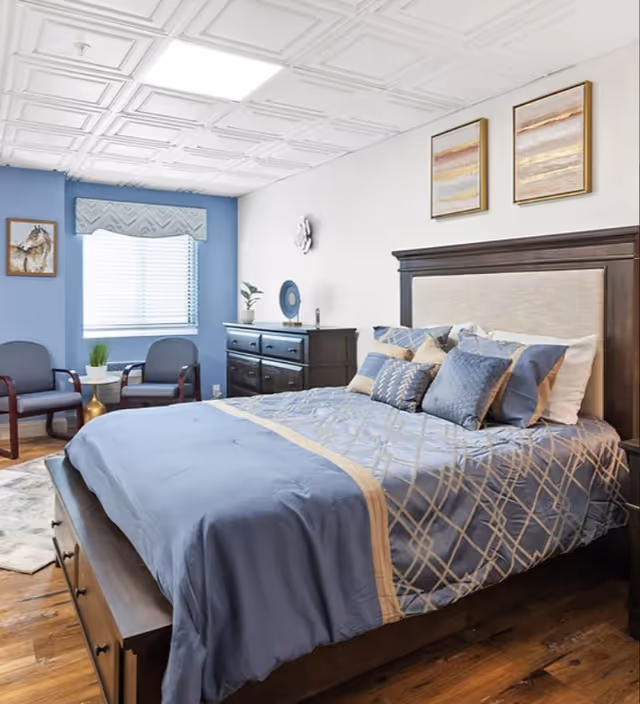 Well-lit bedroom with a blue-dressed bed, wooden headboard and dresser, two chairs by a window, and framed wall art.