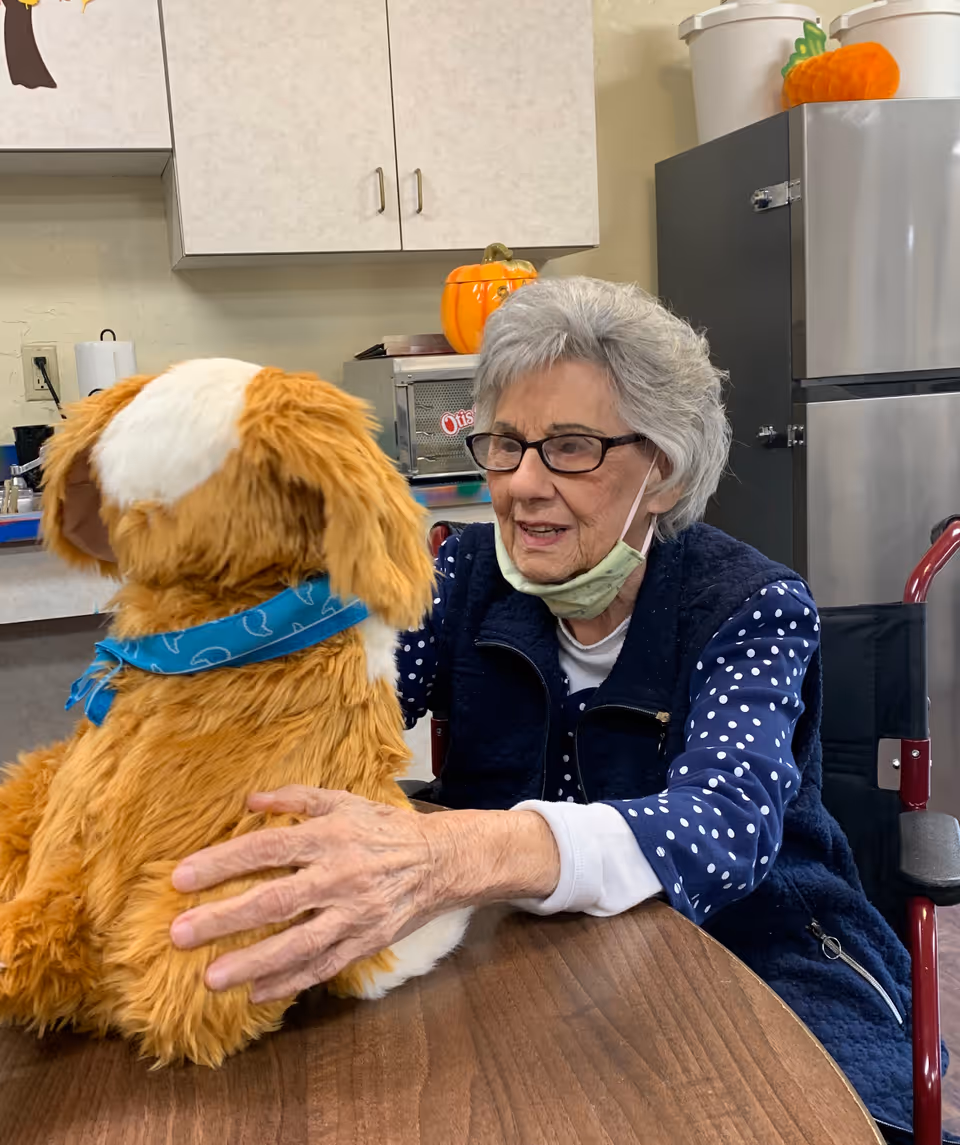 An elderly woman wearing glasses and a face mask pulled down under her chin is sitting at a table in a room with cabinets and a refrigerator. She is interacting with a large, plush dog toy that has a blue bandana around its neck.