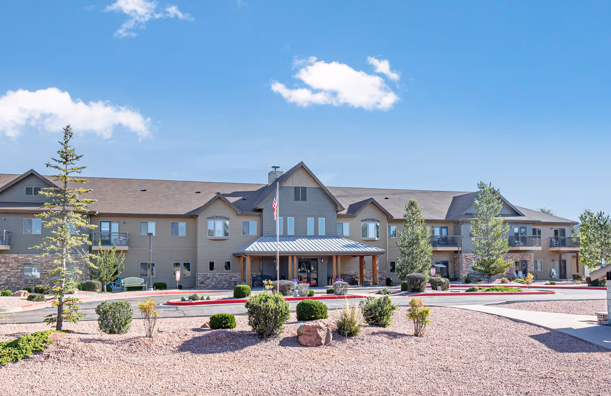 Front exterior view of Majestic Rim Retirement Living facility with a two-story building, landscaped bushes, trees, and a clear blue sky with a few clouds.