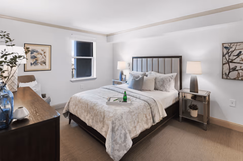 A neatly made bed with a padded headboard in a bright bedroom. The bed has white and light gray bedding with multiple pillows. There are two bedside tables with lamps on each side of the bed. A framed artwork hangs on the wall to the right, and another framed picture is on the left wall near a window. A wooden dresser with decorative items, including a blue vase and a small plant, is visible in the foreground. The room has beige carpeting and white walls.