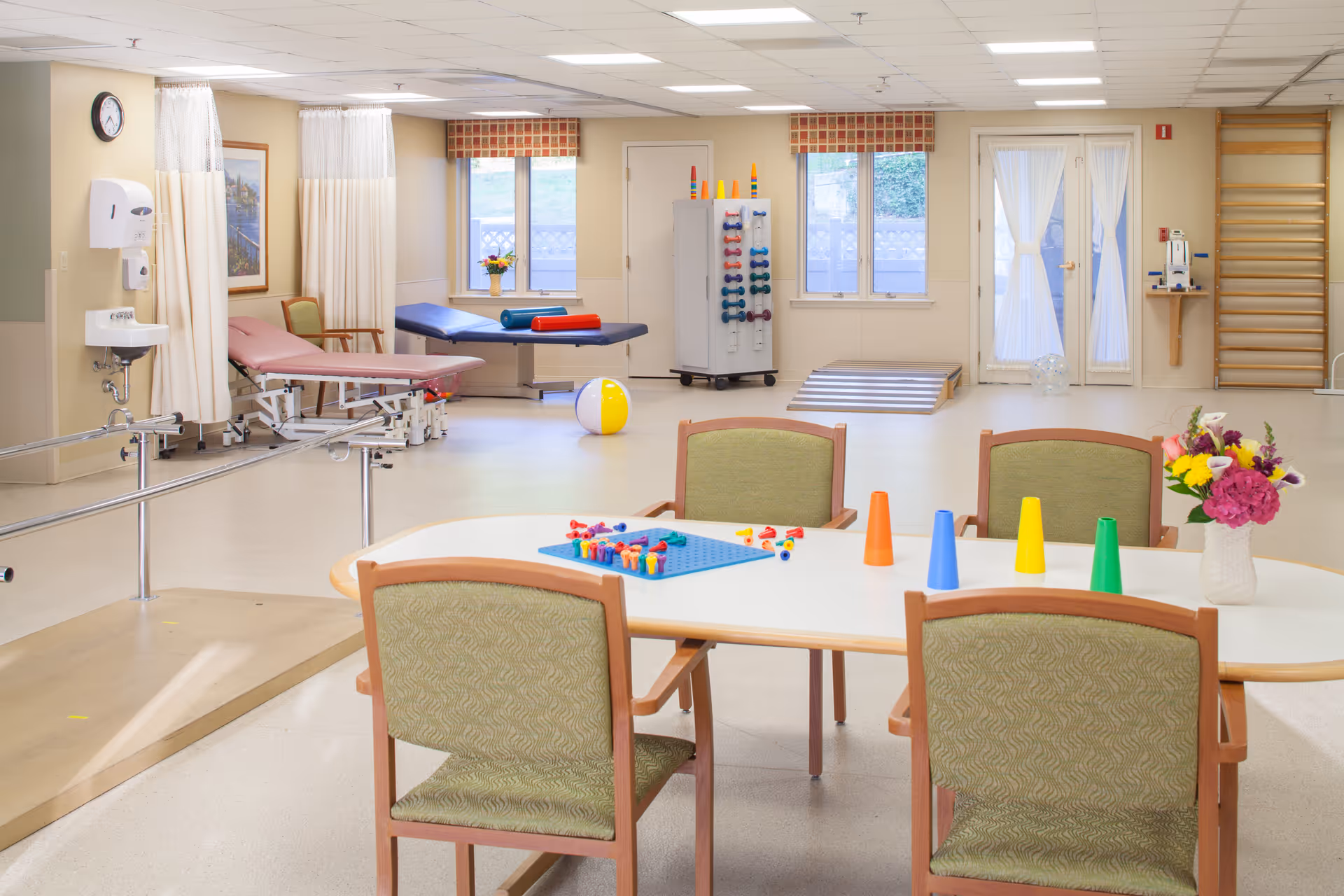 Bright multipurpose rehabilitation/activity room with a table and chairs in the foreground and therapy tables, exercise equipment, and colorful cones and toys in the background.