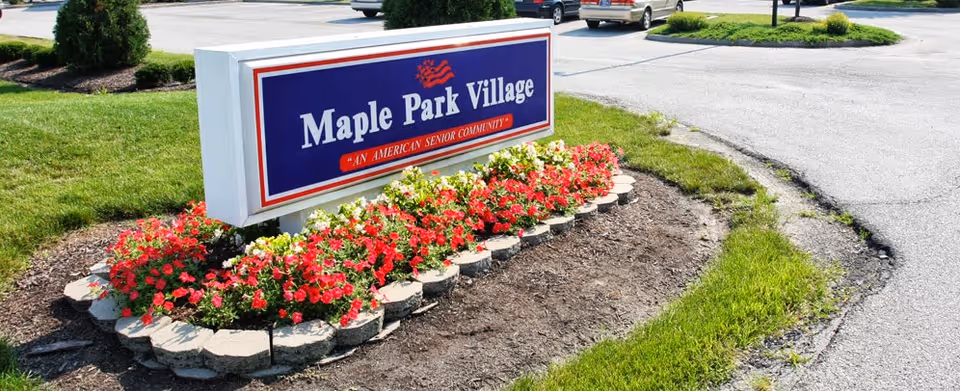 A roadside sign for Maple Park Village, an American senior community, surrounded by a flower bed with red and white flowers and bordered by stone edging. The sign is positioned near a paved road with grass and some parked cars visible in the background.