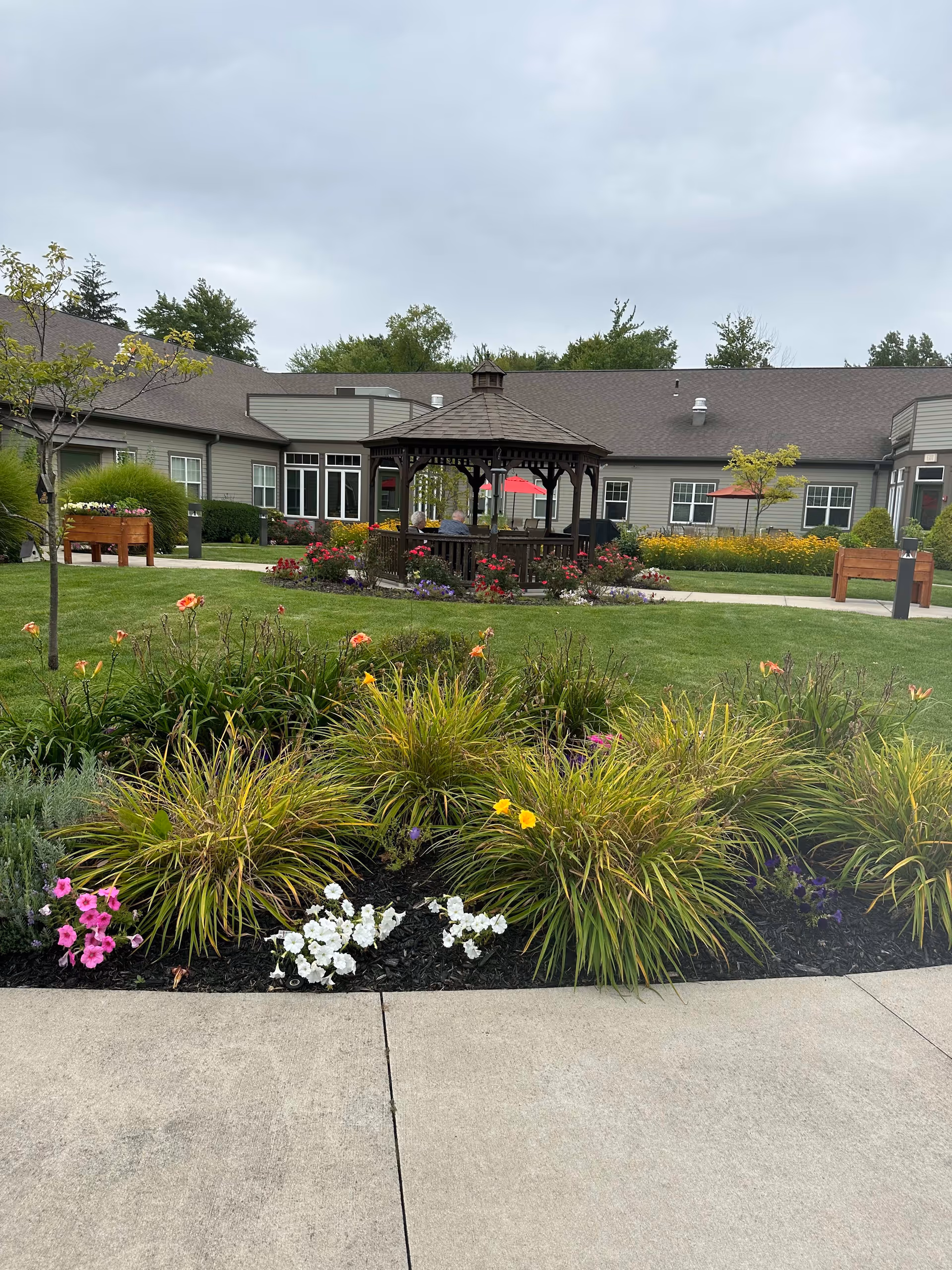 Outdoor garden area at North Woods Village at Edison Lakes featuring a wooden gazebo surrounded by colorful flowers and green shrubs, with a building in the background under a cloudy sky.
