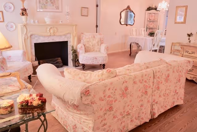 A pastel floral-upholstered living room with a sofa, armchairs, fireplace, and a dining table visible in the background.
