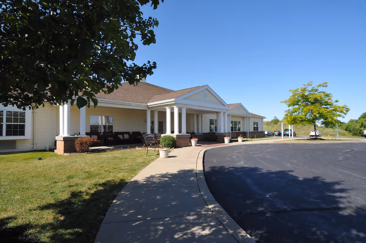 Exterior view of a single-story building with a covered entrance supported by white columns. There is a curved driveway and a sidewalk leading to the entrance. The building is surrounded by green grass, a few trees, and several parked cars under a clear blue sky.