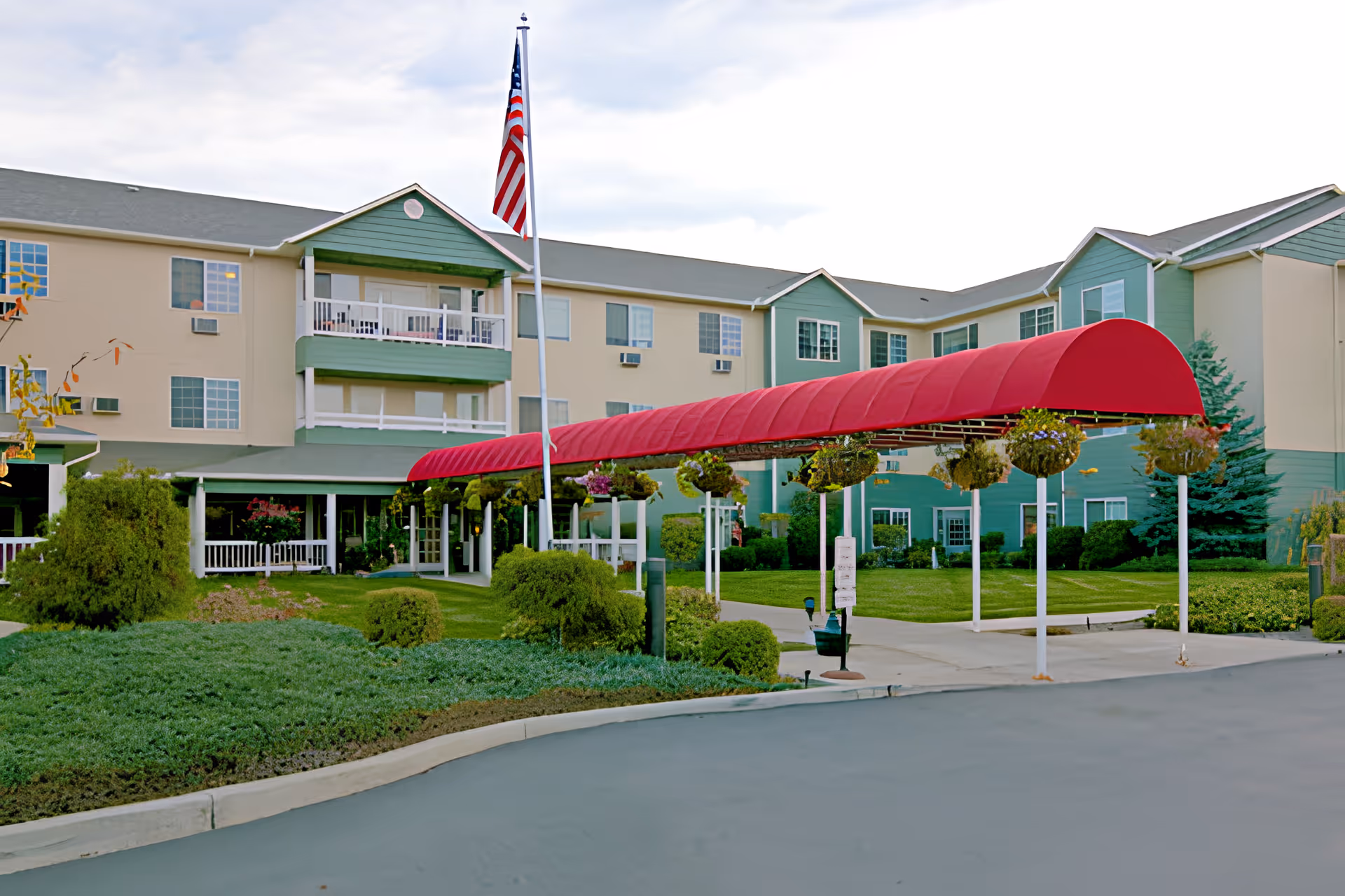 Exterior view of a senior living facility with a three-story building featuring balconies and multiple windows. A red covered walkway extends from the entrance to the driveway, with hanging flower baskets along the walkway. An American flag is displayed on a flagpole near the entrance, surrounded by well-maintained greenery and bushes.