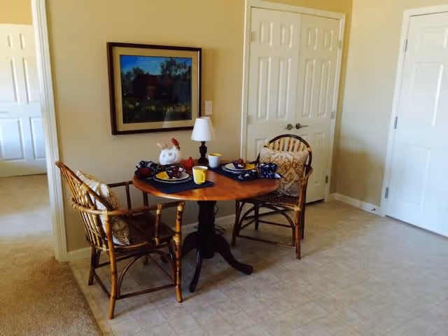 A small dining area with a round wooden table set for two, featuring plates with food, cups, and a small lamp. Two wicker chairs with cushions are placed around the table. The room has beige walls, white double doors, and a framed picture hanging on the wall.