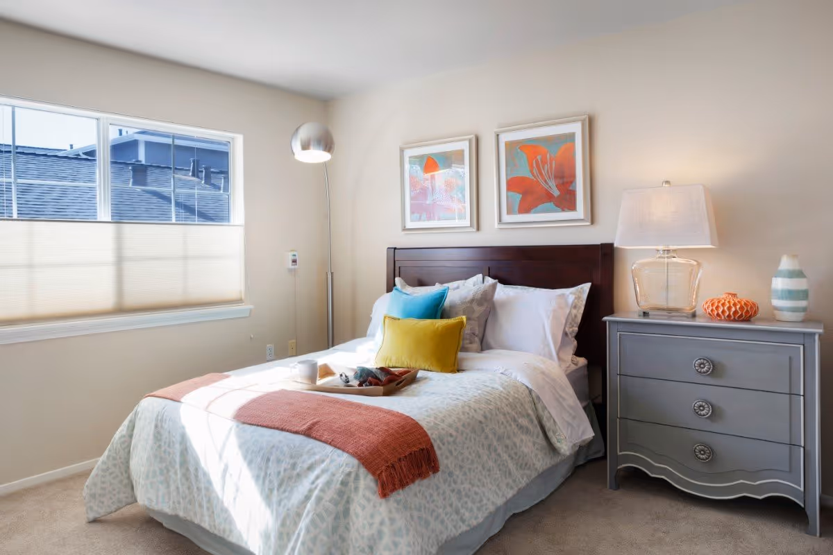 A bright bedroom with a neatly made bed featuring white and patterned bedding, colorful pillows, and a coral throw blanket. There is a gray nightstand with a lamp, decorative items, and two framed floral artworks hanging above the dark wooden headboard. A window with a beige shade allows natural light to fill the room.