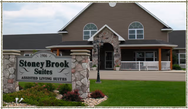 Front exterior view of Stoney Brook Suites Assisted Living building with a stone and wood facade, a sign in front displaying the facility name, a green lawn, and a paved driveway.