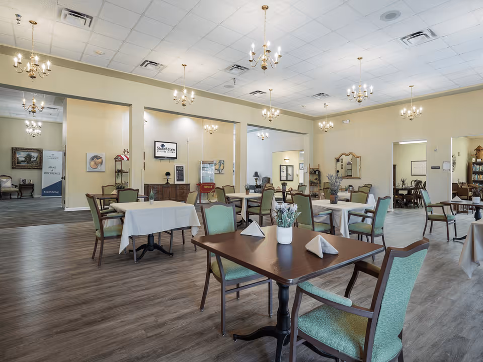 A spacious dining room with multiple tables covered in white tablecloths and set with folded napkins. The room features green cushioned chairs, wooden flooring, beige walls, and several chandeliers hanging from a white ceiling. There are decorative items such as plants and framed pictures on the walls, and a sign that reads 'Stonehaven Assisted Living' is visible in the background.