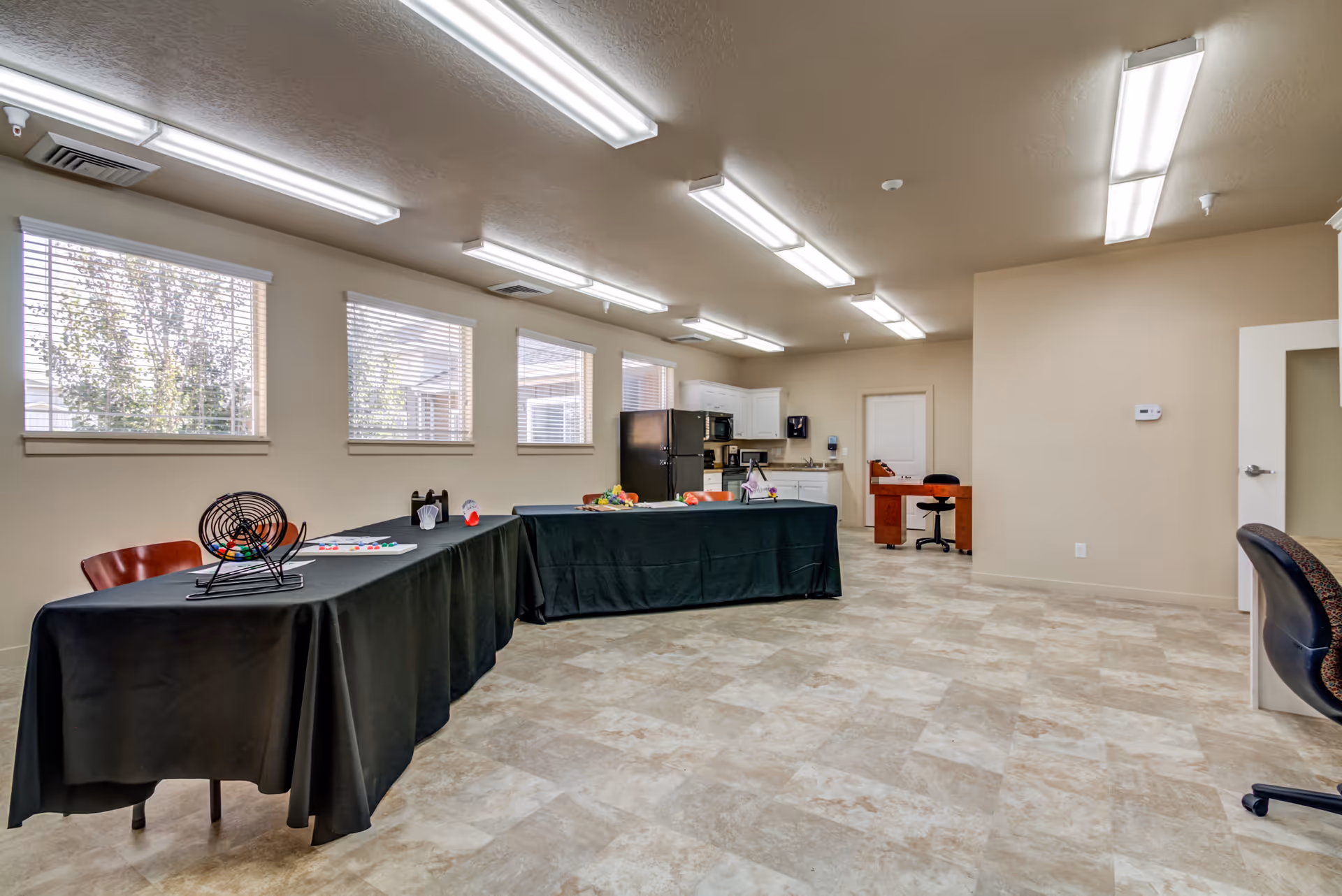 A spacious room with beige walls and tiled floor, featuring two tables covered with black tablecloths arranged in an L shape. On the tables are various items including a bingo cage, bingo cards, and other small objects. The room has several windows with blinds letting in natural light, fluorescent ceiling lights, a black refrigerator, white kitchen cabinets, a microwave, and a small desk with a chair in the background.