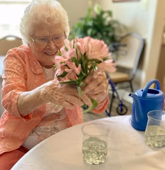 An elderly woman with white hair and glasses is smiling while holding a bouquet of pink flowers. She is seated at a table with a white tablecloth, which has two clear plastic cups with water and a blue watering can on it. In the background, there is a walker and some chairs, suggesting an indoor setting in a senior living facility.