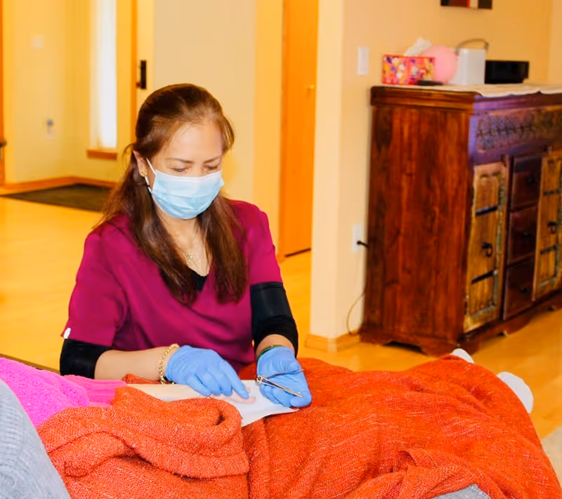 A caregiver wearing a face mask and gloves is attending to a person lying down covered with an orange blanket inside a warm-toned room with wooden furniture.
