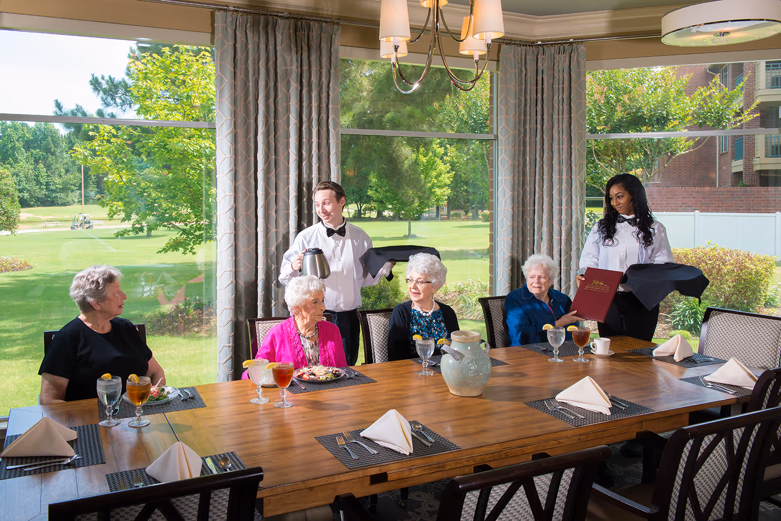Four elderly women seated around a wooden dining table with place settings and drinks, two waitstaff standing nearby serving and holding a menu, large windows with curtains showing a green outdoor view in the background.