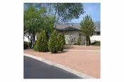 A single-story building with a gray roof and beige walls surrounded by trees and shrubs, with a gravel-covered front yard and a paved driveway or street in front.