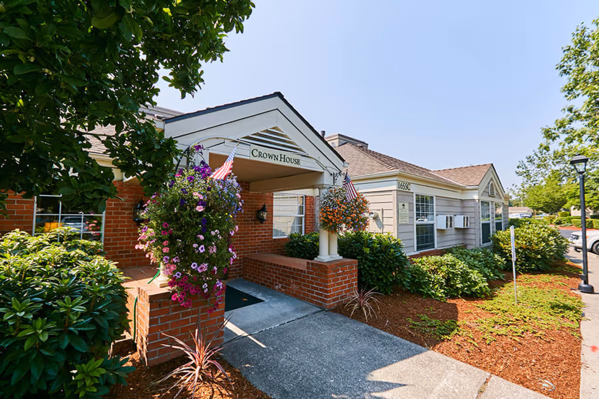 Exterior view of a single-story building entrance labeled 'Crown House' with hanging flower baskets and American flags. The building has brick and siding walls, surrounded by green shrubs and trees under a clear blue sky.
