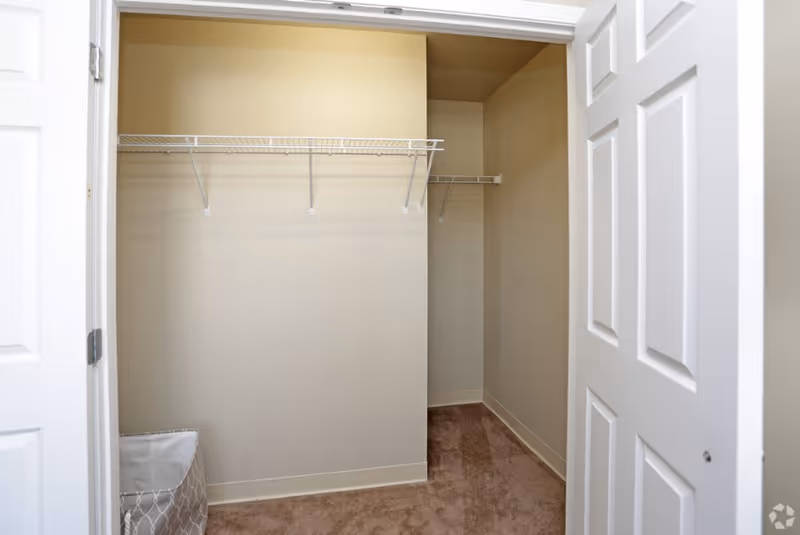 Empty walk-in closet with beige walls, carpeted floor, white wire shelving on two walls, and white double doors partially open.