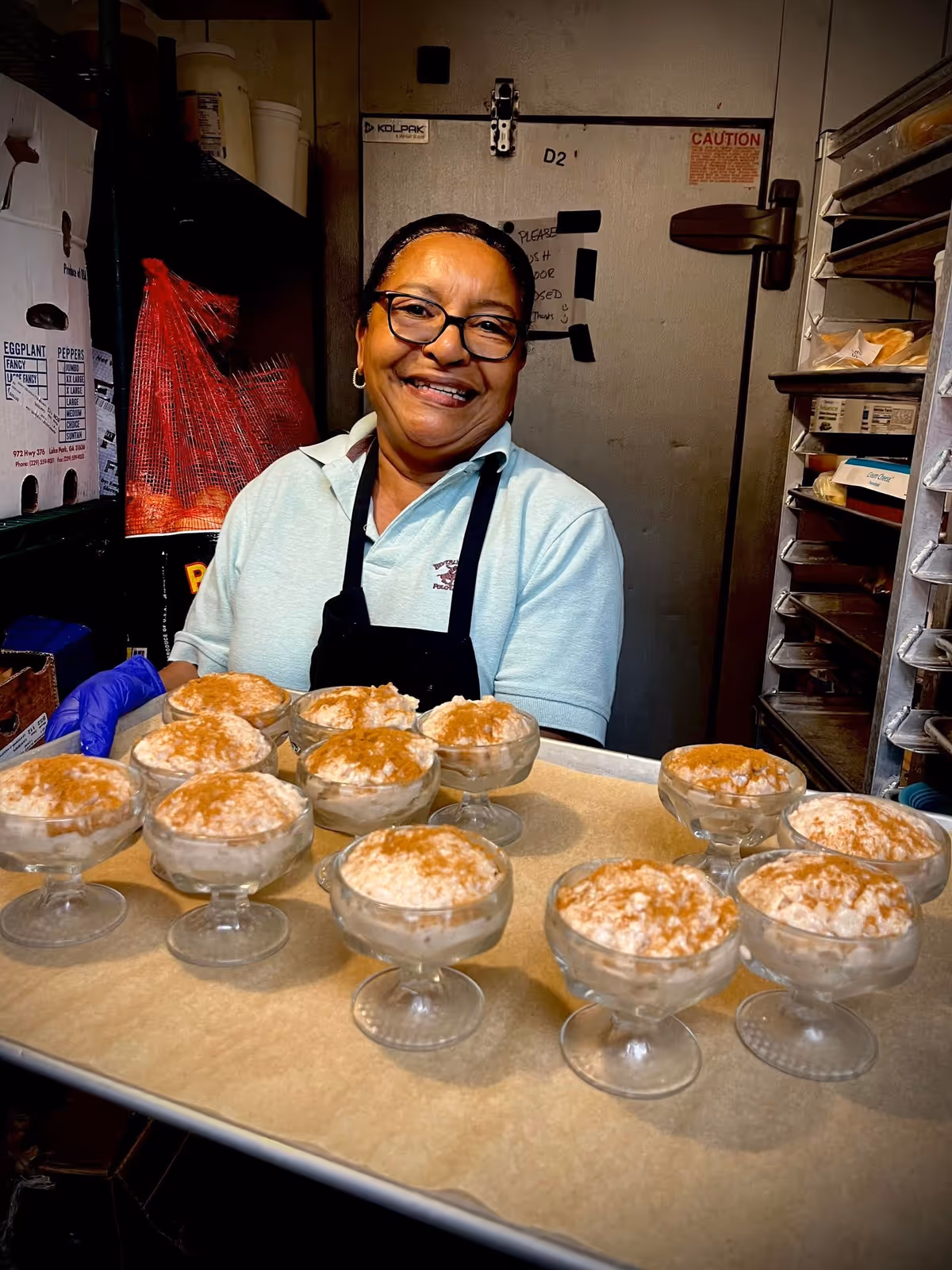A smiling woman wearing glasses, a light blue shirt, and a black apron holds a tray with several glass dessert cups filled with a creamy dessert topped with cinnamon. She is standing in a kitchen or food preparation area with shelves and a refrigerator door in the background.
