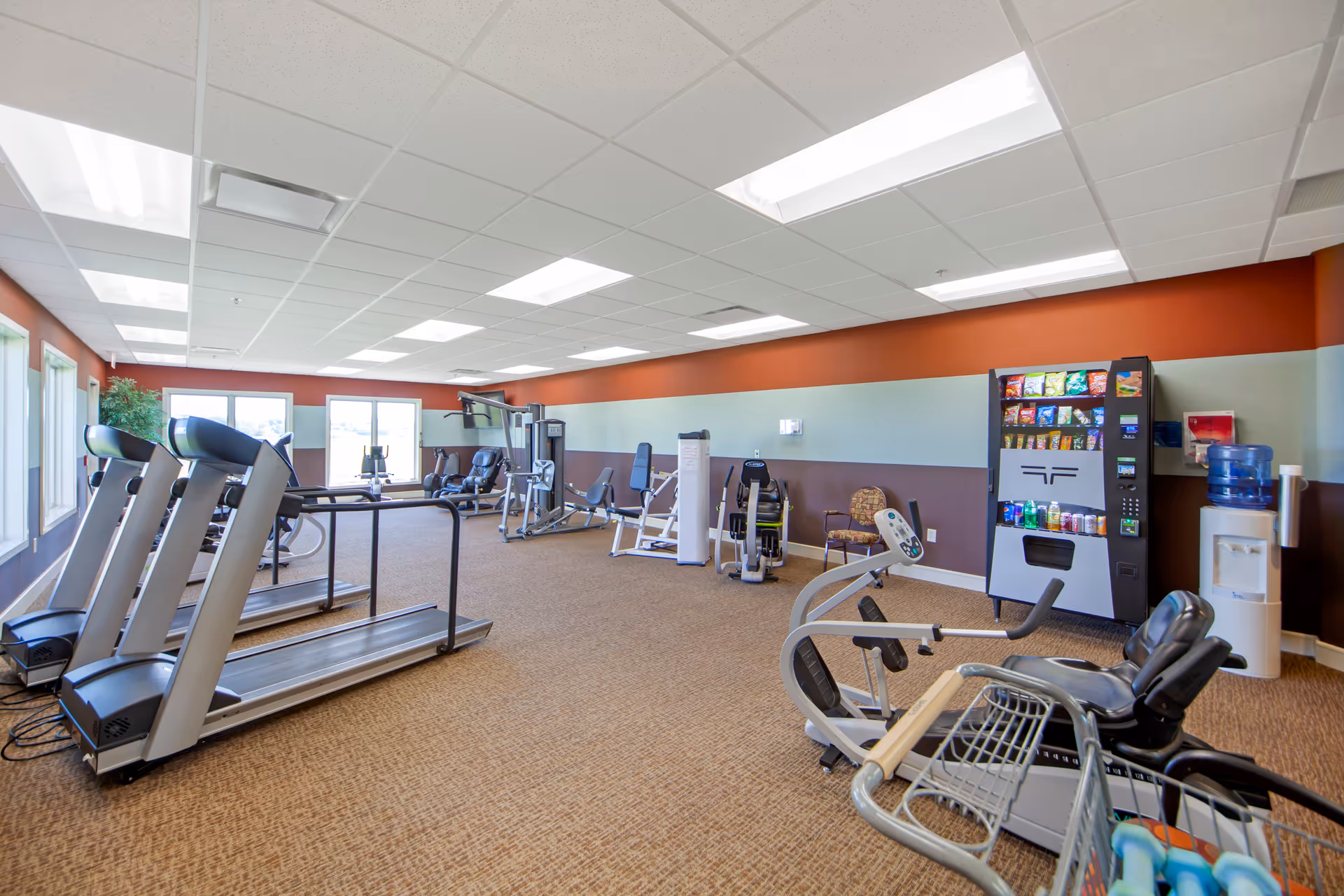 A spacious fitness room with various exercise equipment including treadmills, stationary bikes, and weight machines. The room has large windows letting in natural light, a vending machine stocked with snacks and drinks, and a water cooler. The walls are painted in a combination of brown, light blue, and orange, and the floor is carpeted.