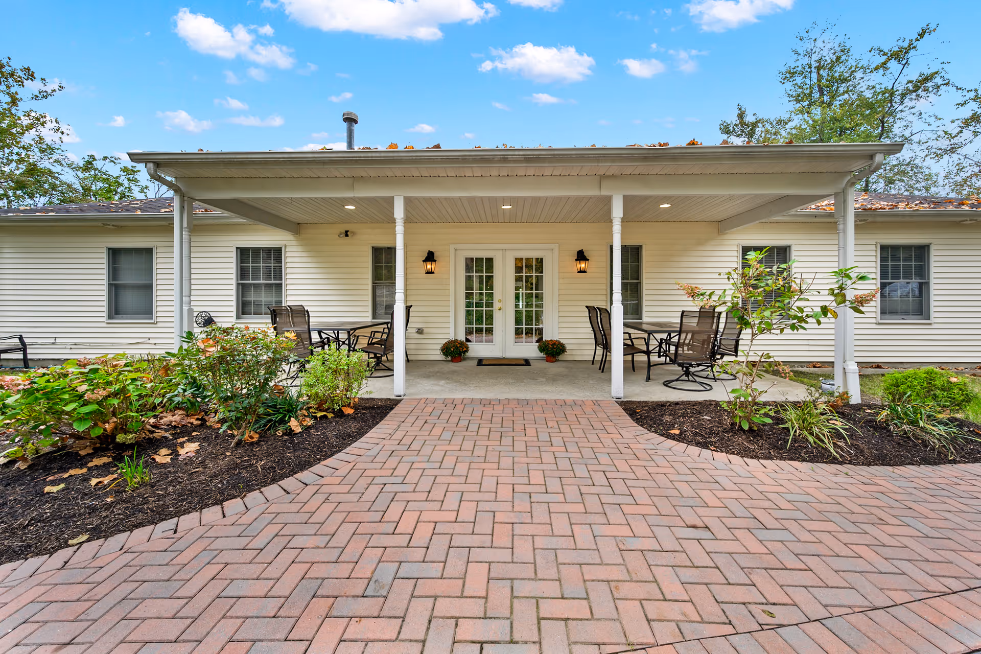 Covered entrance patio of a single-story memory care building with a brick paver walkway, outdoor seating, and double glass doors.