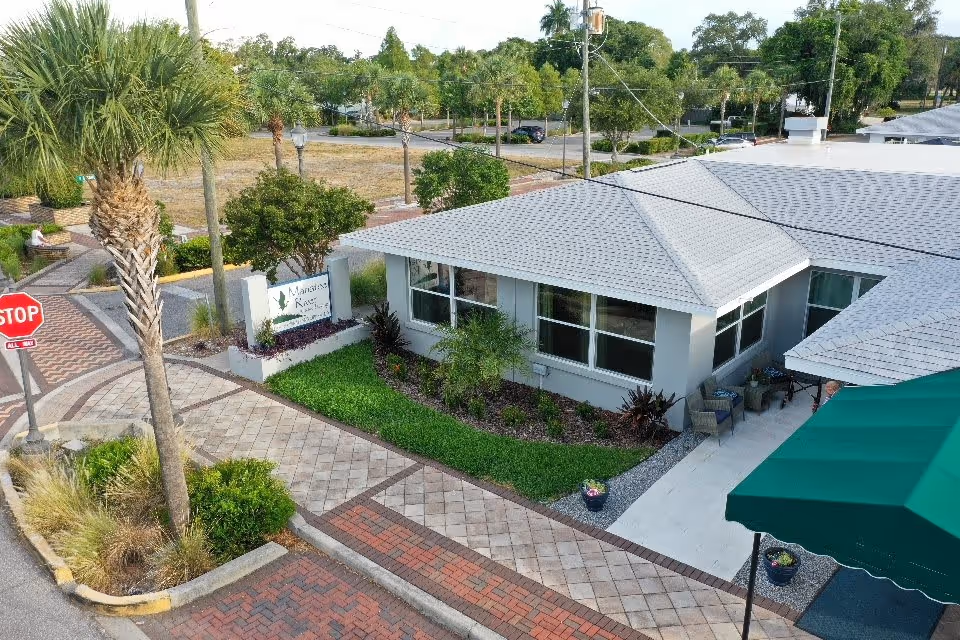 Exterior view of Manatee River Assisted Living facility showing a single-story building with a gray roof, large windows, landscaped greenery, a paved walkway, a stop sign, and a green awning over an entrance area.