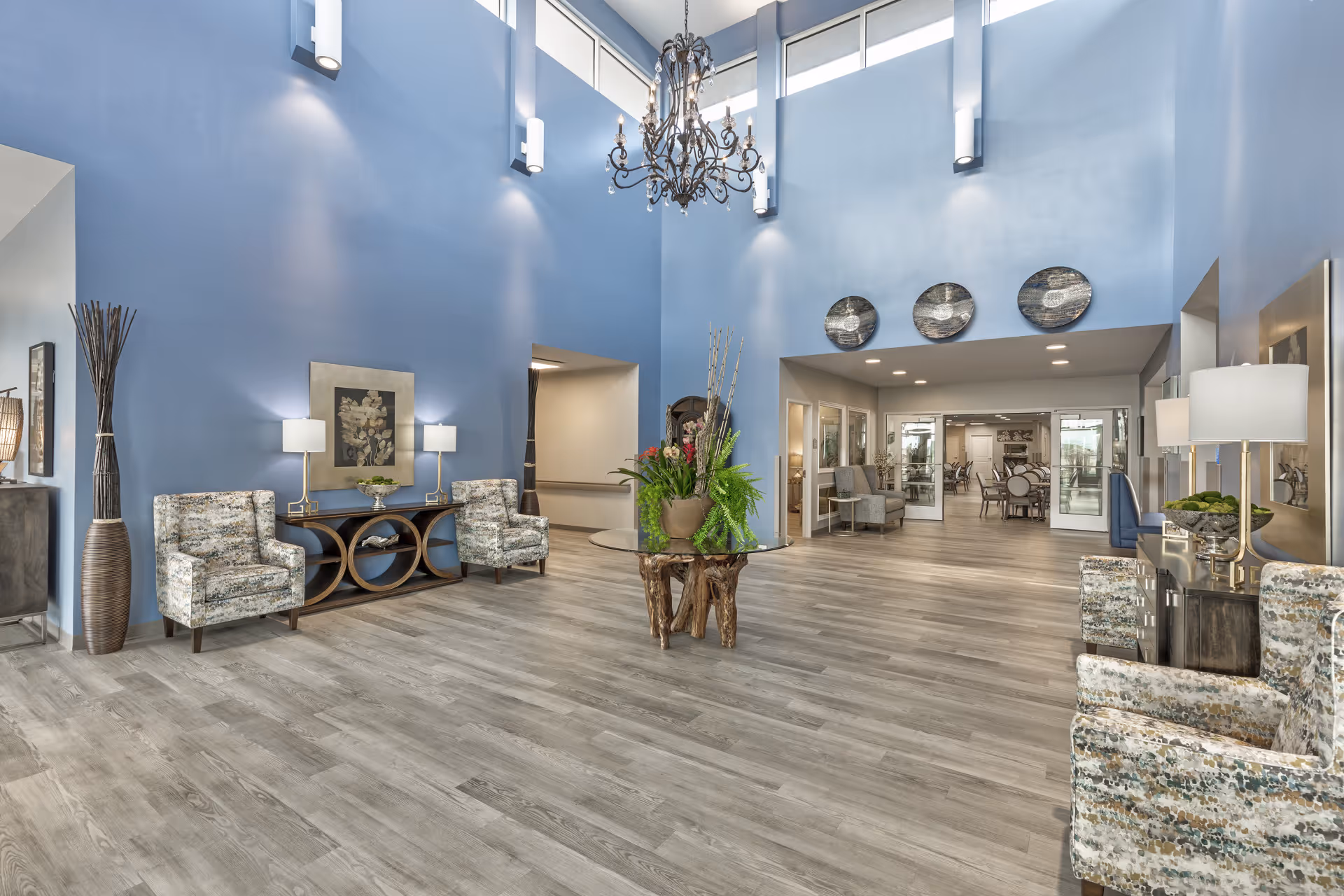 Spacious high-ceilinged lobby with blue walls, seating areas, a central plant display, and a chandelier.