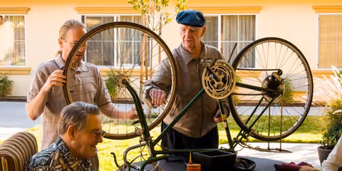 Three elderly men outdoors at Cedar Crest Alzheimer's Special Care Center, gathered around a bicycle that is flipped upside down. One man is holding the bicycle wheel, another is inspecting the bike, and the third is seated nearby, all engaged in a bike repair or maintenance activity. The background shows a building with windows and some greenery.