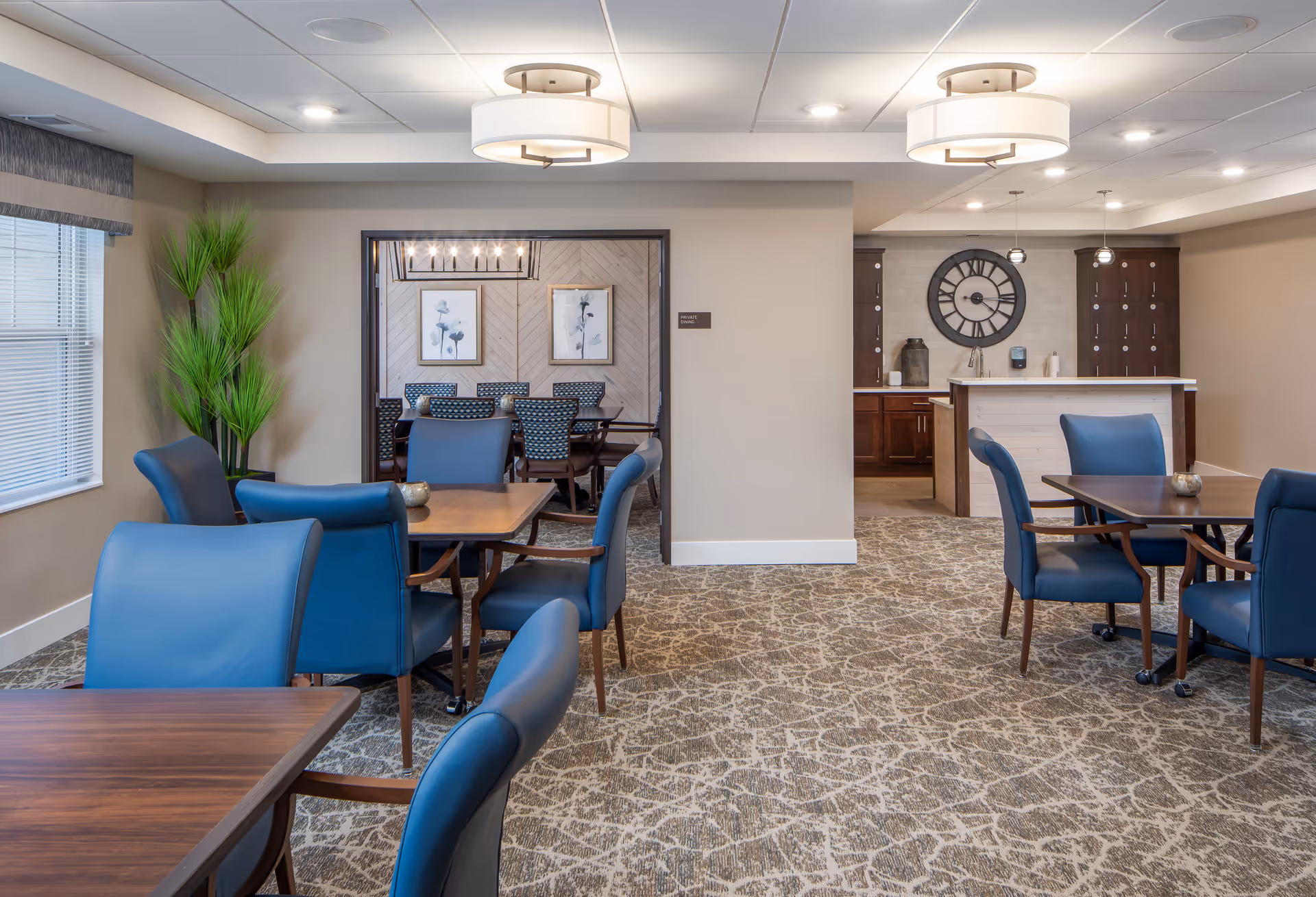A well-lit dining area in a senior living facility with several wooden tables and blue upholstered chairs. In the background, there is a smaller private dining room with patterned chairs and framed floral artwork on the wall. To the right, there is a kitchenette area with a large decorative clock on the wall and cabinets below a countertop. The room has beige walls, a patterned carpet, and modern ceiling lights.