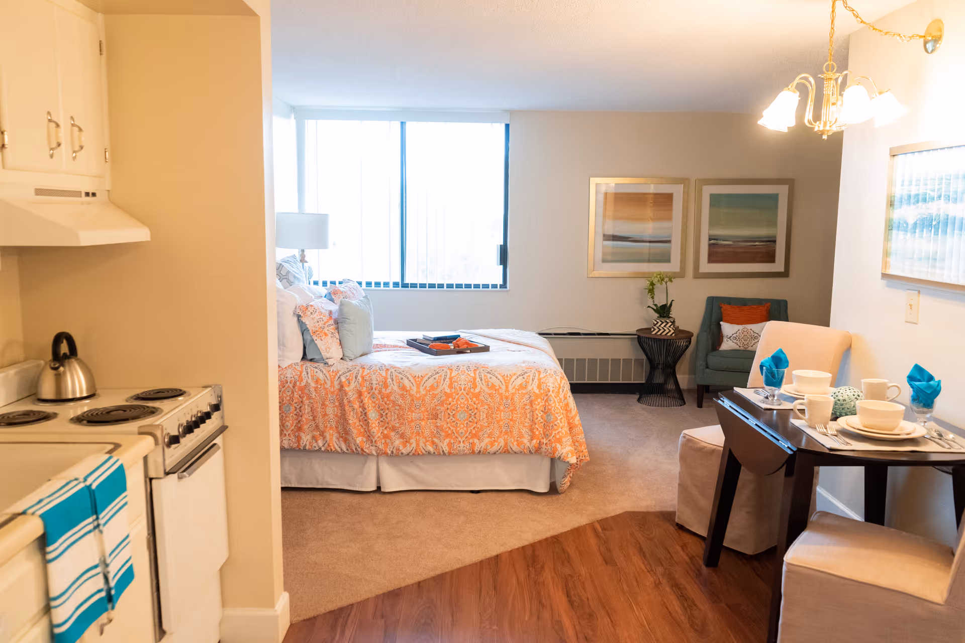 A cozy studio apartment featuring a small kitchen area with a stove and kettle on the left, a bed with orange patterned bedding near a large window in the center, and a dining table set for two with blue napkins on the right. The room also includes a green armchair, framed artwork on the walls, and a small side table with a plant.
