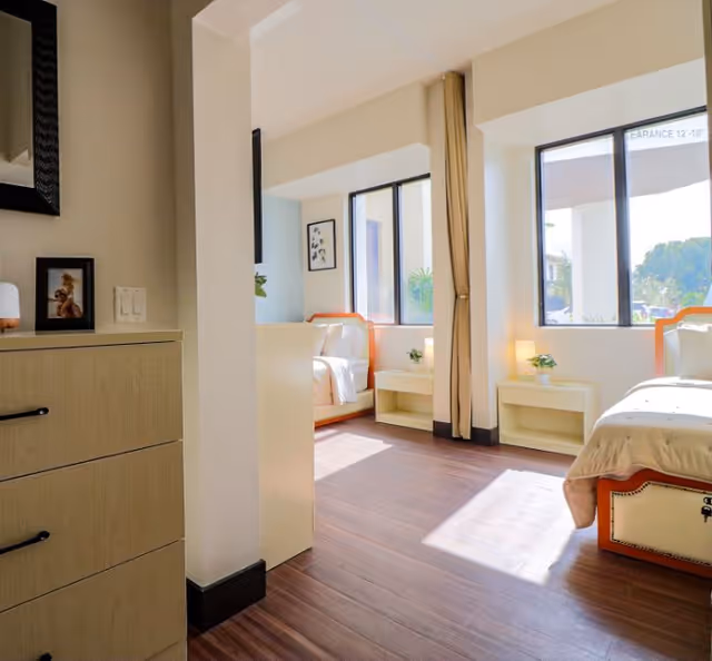 A bright and clean bedroom in a senior living facility with two beds, large windows letting in natural light, light-colored walls, wooden flooring, and small bedside tables with plants.