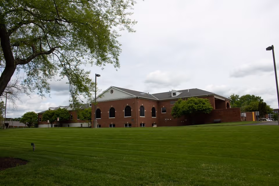 A large brick building with multiple arched windows surrounded by a well-maintained green lawn and trees under a cloudy sky.