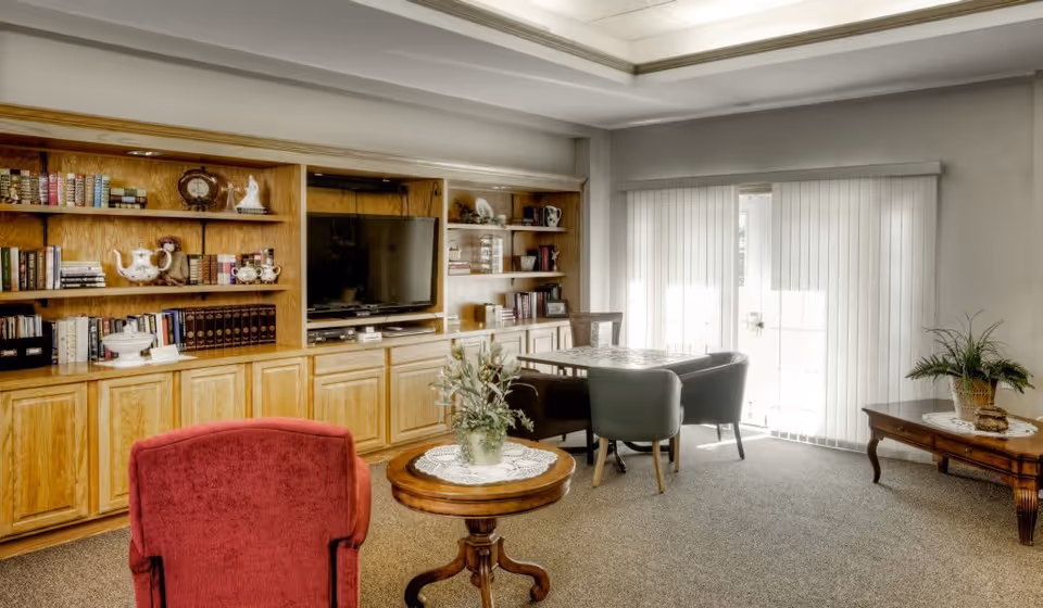 A cozy living room area with built-in wooden shelves filled with books and decorative items, a flat-screen TV, a red upholstered armchair, a round wooden table with a plant centerpiece, a square table with four chairs near sliding glass doors with vertical blinds, and a wooden side table with a plant on it.