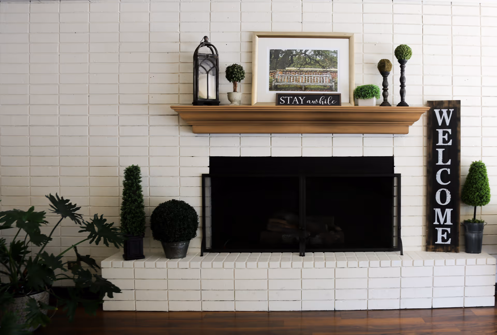 White brick fireplace with a wooden mantel displaying potted plants, framed art, and a vertical "WELCOME" sign.