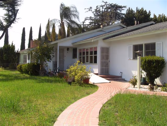Front exterior view of a single-story white building with a sloped roof, a brick pathway leading to the entrance, surrounded by green grass, bushes, and tall trees in the background.