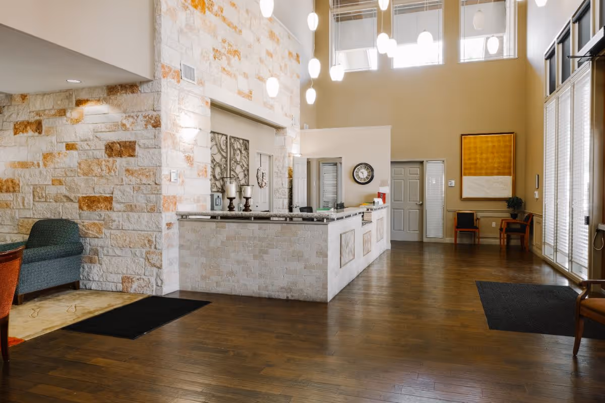 Interior view of a senior living facility reception area with a stone front desk, wooden flooring, high ceilings with hanging pendant lights, and seating areas with chairs along the walls.