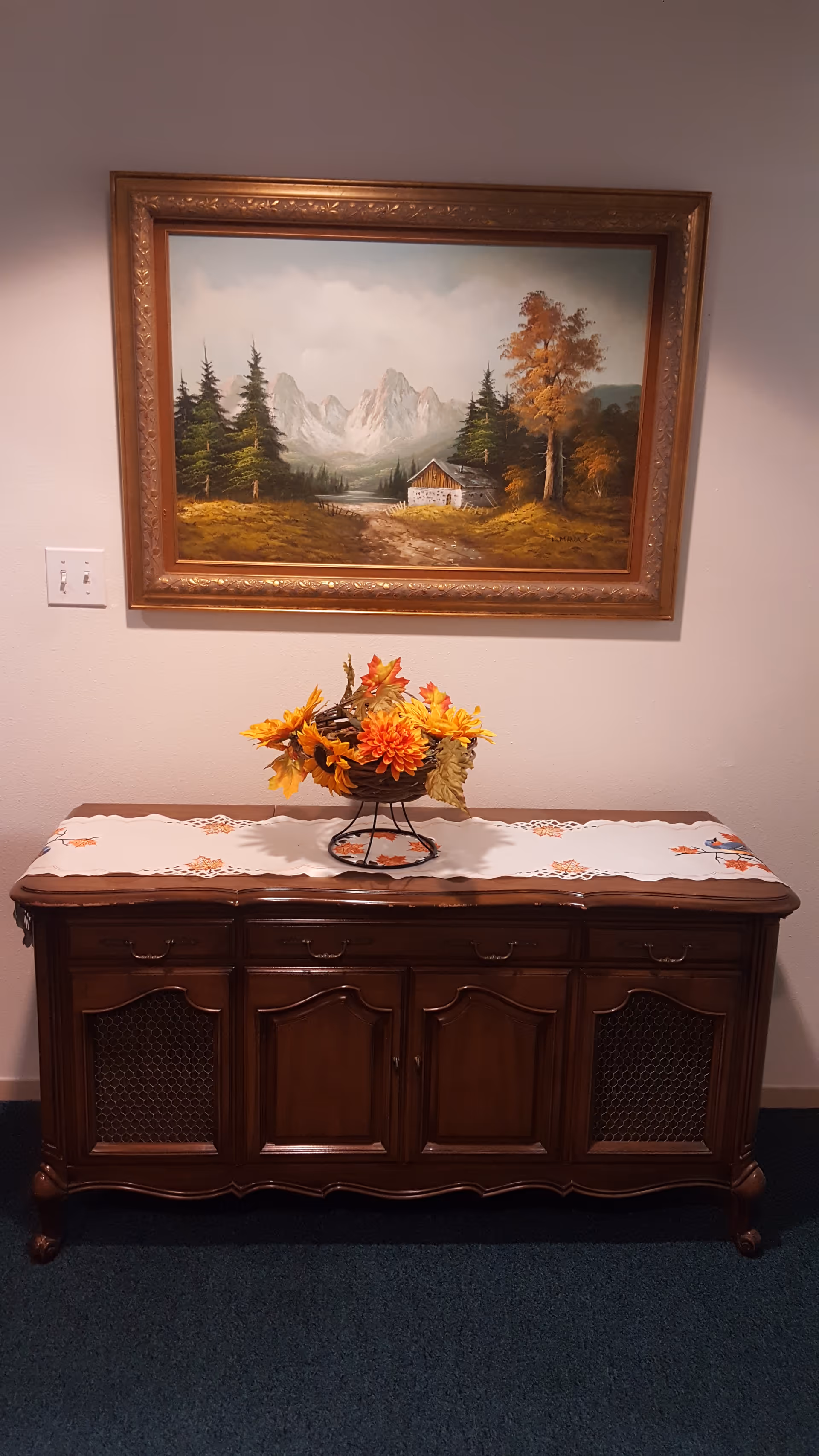 Framed landscape painting above a wooden sideboard with a floral arrangement and decorative runner on a carpeted interior wall.