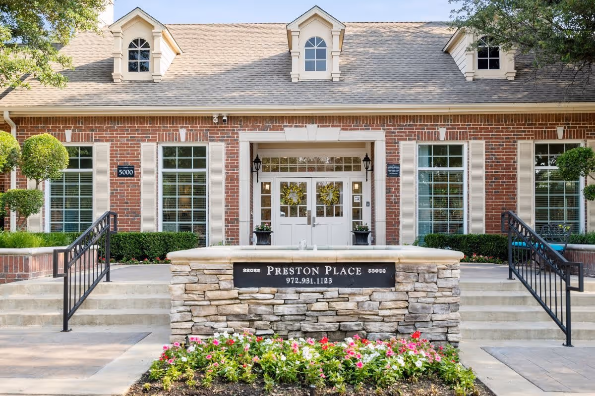 Front exterior view of Preston Place, a brick building with white trim and large windows. There are two staircases with black railings leading up to the entrance, which has double white doors and two black lantern-style lights. In front of the building is a stone sign with the name Preston Place and a phone number, surrounded by a flower bed with pink and white flowers.