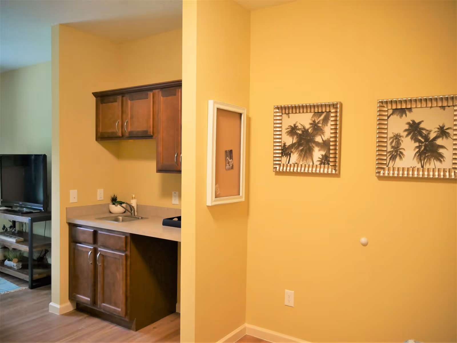 Interior view of a room with yellow walls featuring a small kitchenette with wooden cabinets and a sink. On the right wall, there are two framed pictures of palm trees and a small bulletin board. In the background, a television on a stand is partially visible.