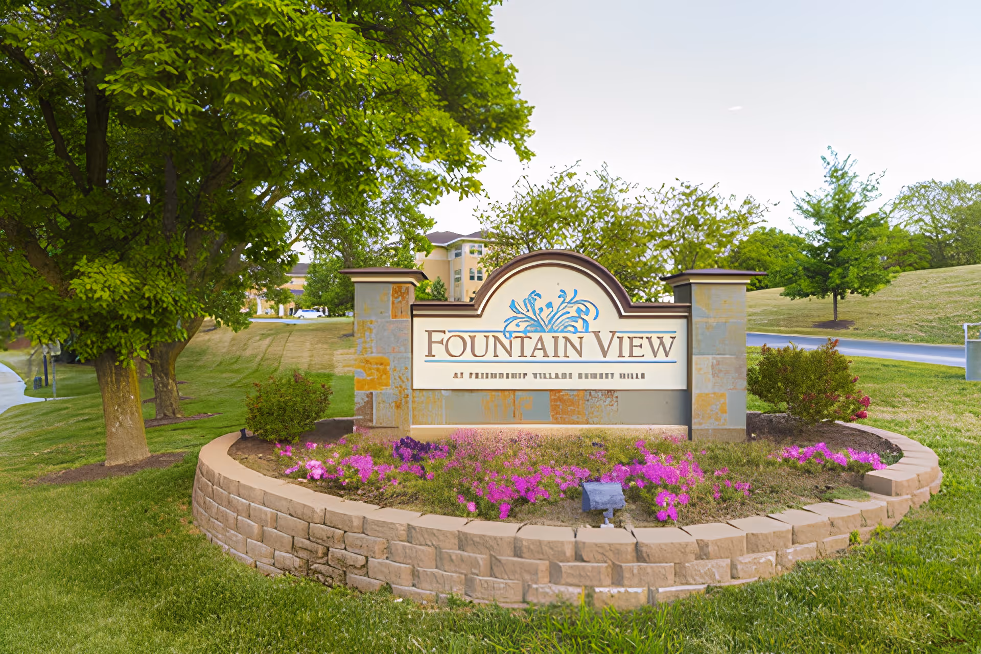 Stone sign for Fountain View Assisted Living surrounded by a curved stone flower bed with pink and purple flowers, green grass, and trees in the background.