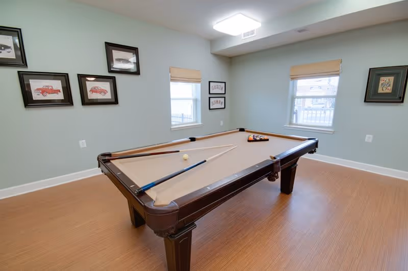 A well-lit room with a pool table in the center, featuring a tan felt surface and two pool cues resting on it. The walls are painted light blue and decorated with framed pictures. Two windows with beige blinds allow natural light into the room, which has a wooden floor.